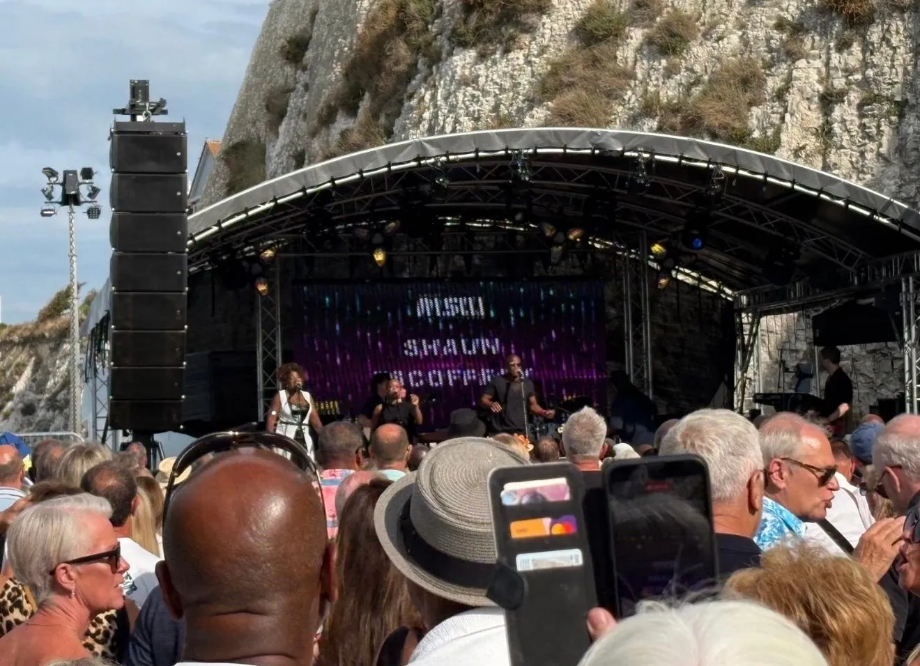 Outdoor stage performance beneath a rocky cliff with an audience in the foreground